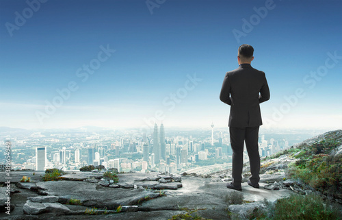 Back view of young businessman standing on mountain top looking at cityscape-Leadership concept