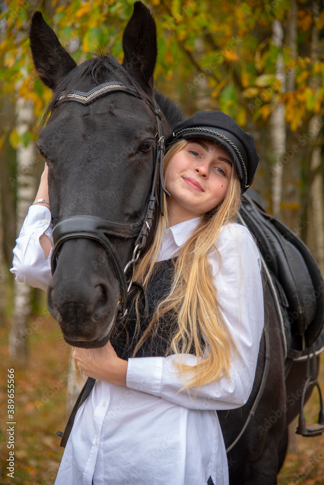 Obraz premium portrait of a girl with blonde hair , wearing a cap, and a Bay horse on the background of a forest. 
