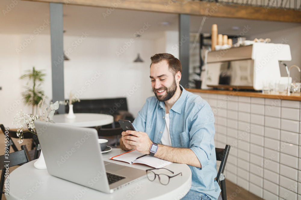 Smiling young man sitting at table in coffee shop cafe working studying on laptop pc computer using cell phone relaxing in restaurant during free time indoor. Freelance mobile office business concept. Smiling young man sitting at table in coffee shop cafe working studying on laptop pc computer using cell phone relaxing in restaurant during free time indoor. Freelance mobile office business concept.