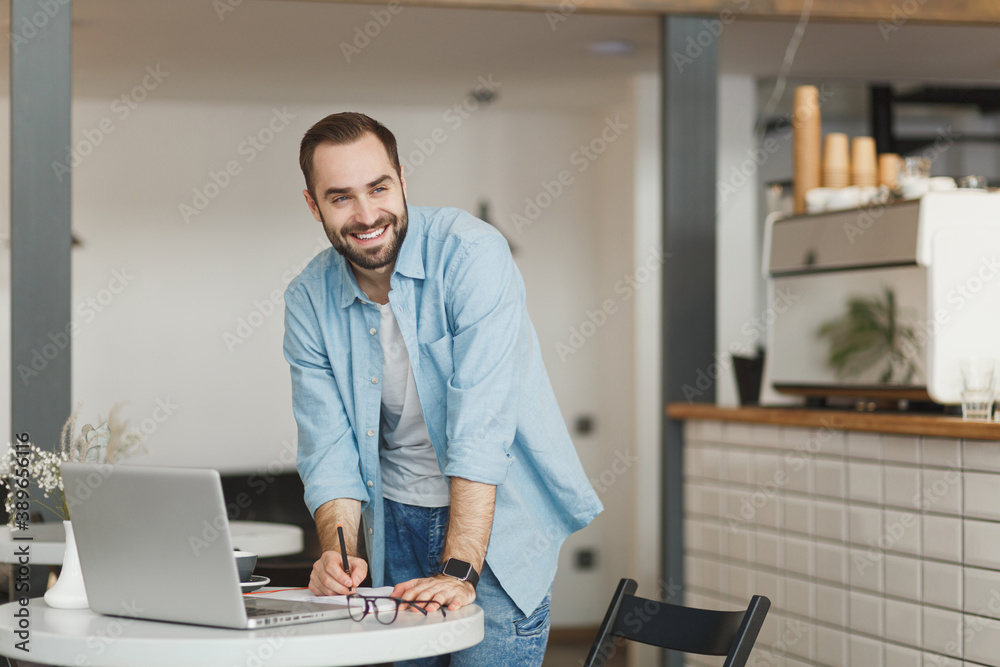 Smiling cheerful young man standing alone near table in coffee shop cafe restaurant indoors working or studying on laptop pc computer writing in notebook. Freelance mobile office business concept.
