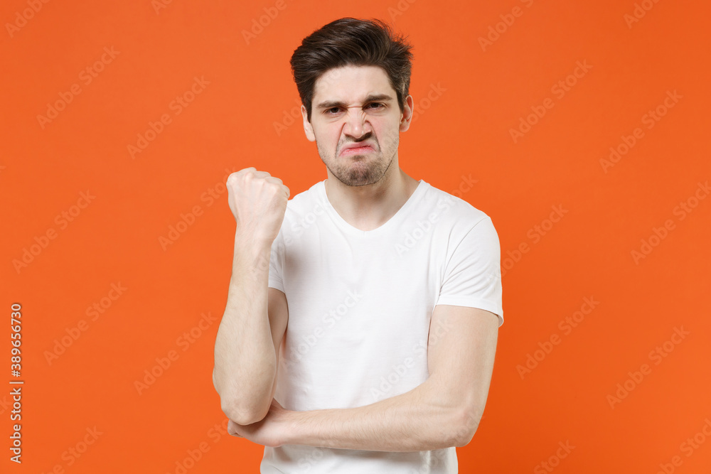 Displeased dissatisfied aggressive young man wearing basic casual empty blank white t-shirt standing clenching fist looking camera isolated on bright orange colour wall background, studio portrait. Displeased dissatisfied aggressive young man wearing basic casual empty blank white t-shirt standing clenching fist looking camera isolated on bright orange colour wall background, studio portrait.