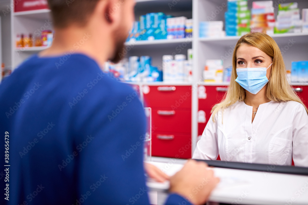 young female apothecary in protective medical mask and young caucasian man customer buying drug at drugstore. medicine, consumerism concept. during coronavirus epidemic