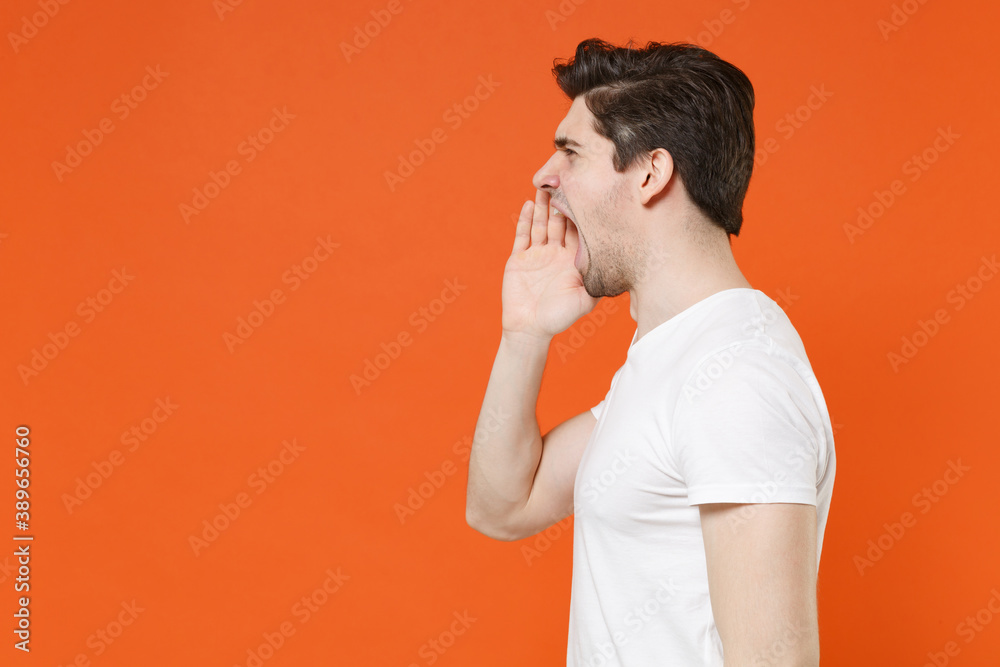 Side view of crazy young man wearing basic casual blank empty white t-shirt standing screaming with hands gesture near mouth looking aside isolated on bright orange colour background, studio portrait. Side view of crazy young man wearing basic casual blank empty white t-shirt standing screaming with hands gesture near mouth looking aside isolated on bright orange colour background, studio portrait.