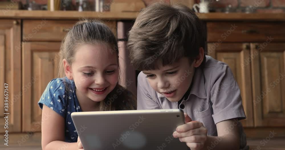 Close up little boy and girl lying on wooden warm floor at home kitchen with tablet device, watch cartoons, online video. Children development using modern technology, addicted kids, bad habit concept Close up little boy and girl lying on wooden warm floor at home kitchen with tablet device, watch cartoons, online video. Children development using modern technology, addicted kids, bad habit concept