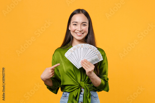 Принти на полотні Smiling funny young brunette asian woman wearing basic green shirt standing pointing index finger on fan of cash money in dollar banknotes isolated on bright yellow colour background, studio portrait