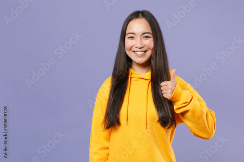 Smiling beautiful attractive young brunette asian woman 20s wearing basic casual yellow hoodie standing showing thumb up looking camera isolated on pastel violet colour background studio portrait.