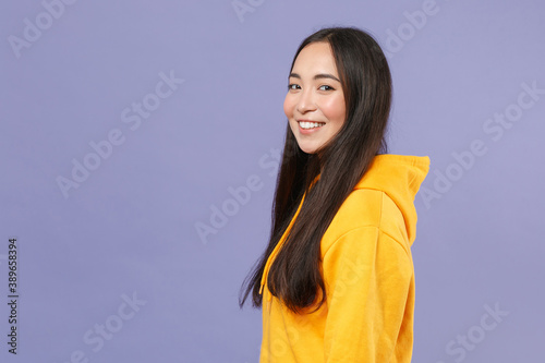 Side view of smiling pretty young brunette asian woman wearing casual basic yellow hoodie standing holding hands crossed looking camera isolated on pastel violet colour background studio portrait.