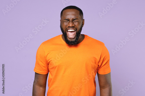 Shocked crazy frustrated young african american man wearing basic casual blank empty orange t-shirt standing keeping eyes closed screaming isolated on pastel violet colour background studio portrait.