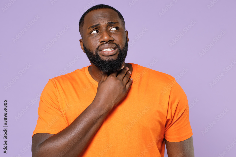 Sick upset young african american man wearing basic casual empty blank orange t-shirt standing feeling pain ache, put hands on throat neck isolated on pastel violet colour background studio portrait. Sick upset young african american man wearing basic casual empty blank orange t-shirt standing feeling pain ache, put hands on throat neck isolated on pastel violet colour background studio portrait.