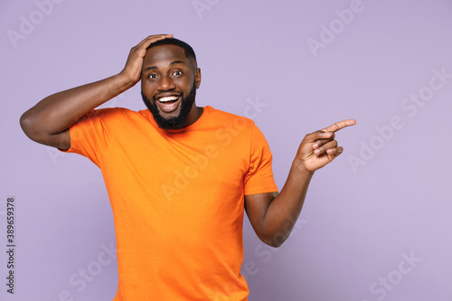Surprised amazed young african american man 20s in basic casual orange t-shirt pointing index finger aside on mock up copy space put hand on head isolated on pastel violet background studio portrait.