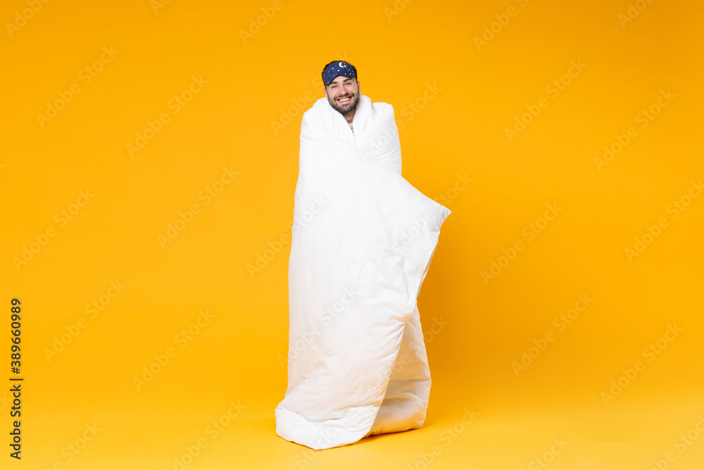 Full length of smiling young bearded man in pajamas home wear sleep mask standing wrapping in blanket isolated on bright yellow colour background studio portrait. Relax good mood lifestyle concept. Full length of smiling young bearded man in pajamas home wear sleep mask standing wrapping in blanket isolated on bright yellow colour background studio portrait. Relax good mood lifestyle concept.