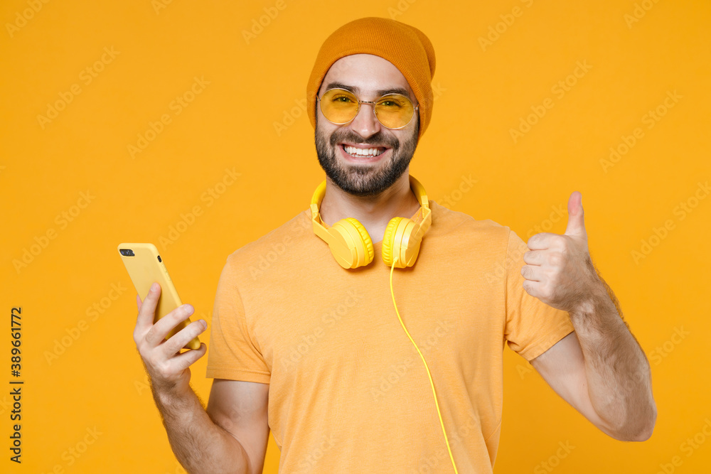 Smiling young bearded man 20s in basic casual t-shirt headphones eyeglasses hat standing using mobile cell phone typing sms message showing thumb up isolated on yellow background, studio portrait. Smiling young bearded man 20s in basic casual t-shirt headphones eyeglasses hat standing using mobile cell phone typing sms message showing thumb up isolated on yellow background, studio portrait.