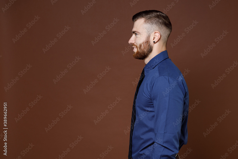 Side view of smiling confident bearded young business man wearing blue shirt tie standing looking aside isolated on brown colour background studio portrait. Achievement career wealth business concept. Side view of smiling confident bearded young business man wearing blue shirt tie standing looking aside isolated on brown colour background studio portrait. Achievement career wealth business concept.