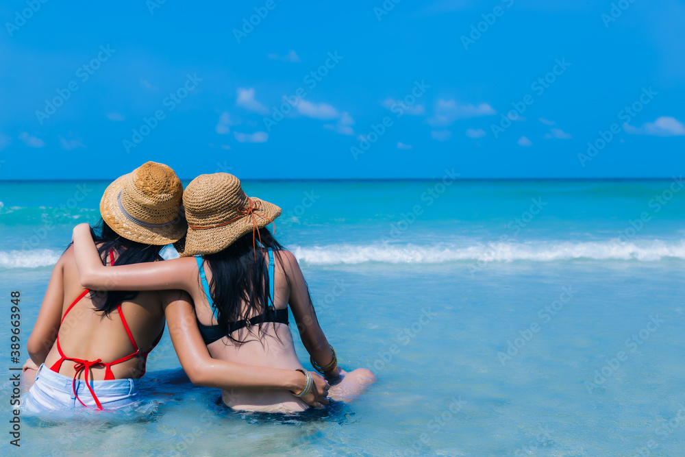 Beautiful young women enjoying on beach and look at nice sea, friend sitting and arms around neck, hug each other on tropical beach in summer vacation time. Traveler girls wear bikini and straw hats
