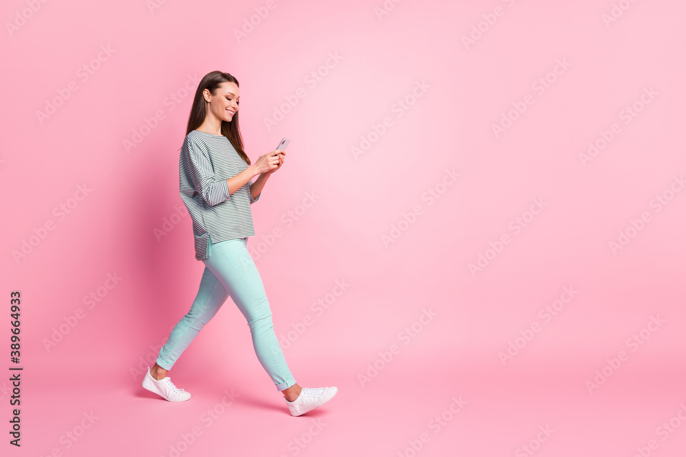 Full size profile photo of optimistic cute young woman going look telephone wear blue jeans sneakers shirt isolated on pink color background