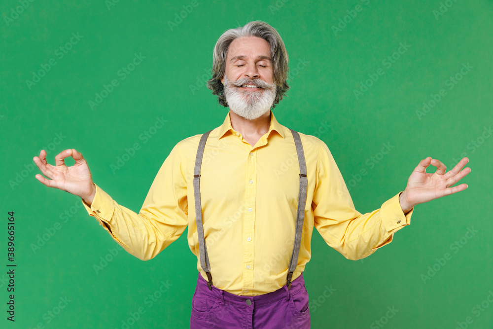 Smiling elderly gray-haired mustache bearded man in yellow shirt suspenders hold hands in yoga gesture, relaxing meditating, trying to calm down isolated on green colour background, studio portrait. Smiling elderly gray-haired mustache bearded man in yellow shirt suspenders hold hands in yoga gesture, relaxing meditating, trying to calm down isolated on green colour background, studio portrait.