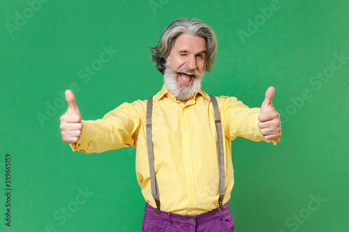 Blinking funny elderly gray-haired mustache bearded man wearing casual yellow shirt suspenders standing showing thumbs up looking camera isolated on bright green colour background, studio portrait. © ViDi Studio