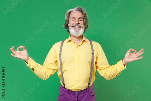 Smiling elderly gray-haired mustache bearded man in yellow shirt suspenders hold hands in yoga gesture, relaxing meditating, trying to calm down isolated on green colour background, studio portrait. © ViDi Studio