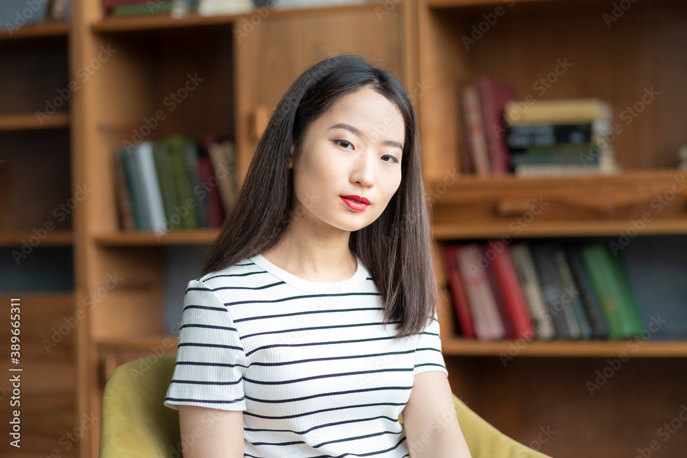 Portrait of beautiful young Asian woman in striped t shirt sitting in chair against background of bookcases in library or business office Portrait of beautiful young Asian woman in striped t shirt sitting in chair against background of bookcases in library or business office