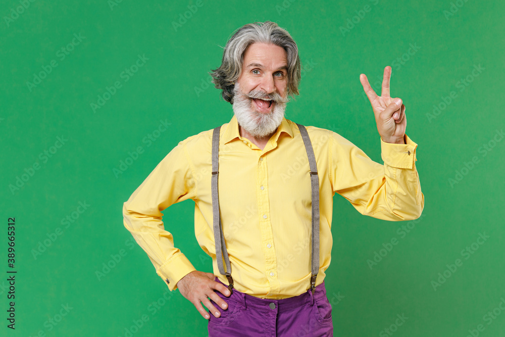 Excited funny elderly gray-haired mustache bearded man wearing basic yellow shirt suspenders standing showing victory sign looking camera isolated on bright green colour background studio portrait.