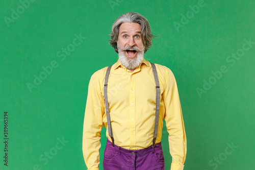 Shocked excited elderly gray-haired mustache bearded man wearing casual yellow shirt suspenders standing keeping mouth open looking camera isolated on bright green colour background, studio portrait. © ViDi Studio