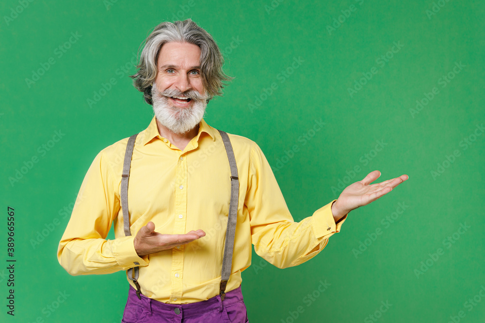 Cheerful funny elderly gray-haired mustache bearded man wearing casual yellow shirt suspenders pointing hands aside on mock up copy space isolated on bright green colour background studio portrait.