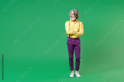 Full length of smiling elderly gray-haired mustache bearded man wearing casual yellow shirt suspenders holding hands crossed looking aside isolated on bright green colour background, studio portrait. © ViDi Studio