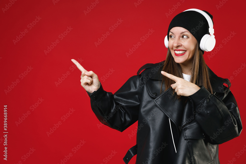 Φωτογραφία Smiling funny young brunette woman 20s in black leather jacket hat standing listening music with headphones pointing index fingers aside isolated on bright red colour wall background studio portrait