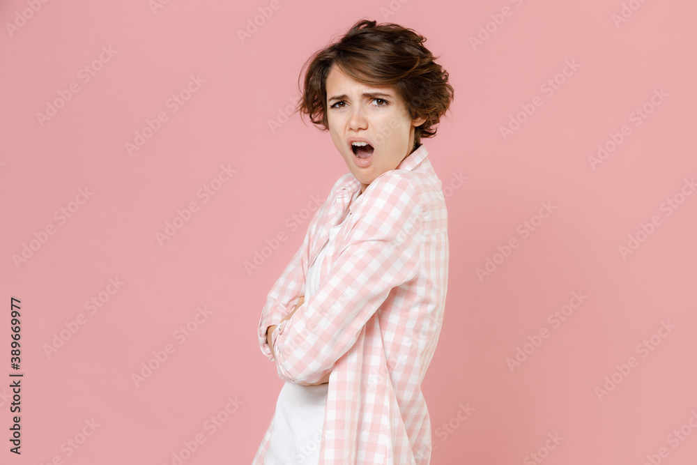 Side view of shocked worried irritated young brunette woman 20s in casual checkered shirt standing holding hands crossed looking camera isolated on pastel pink colour background, studio portrait. Side view of shocked worried irritated young brunette woman 20s in casual checkered shirt standing holding hands crossed looking camera isolated on pastel pink colour background, studio portrait.