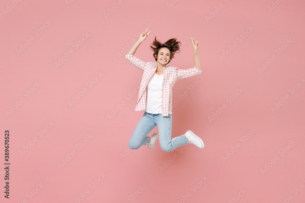 Full length of smiling cheerful funny young brunette woman 20s wearing casual checkered shirt jumping rising hands showing victory sign isolated on pastel pink colour background, studio portrait.