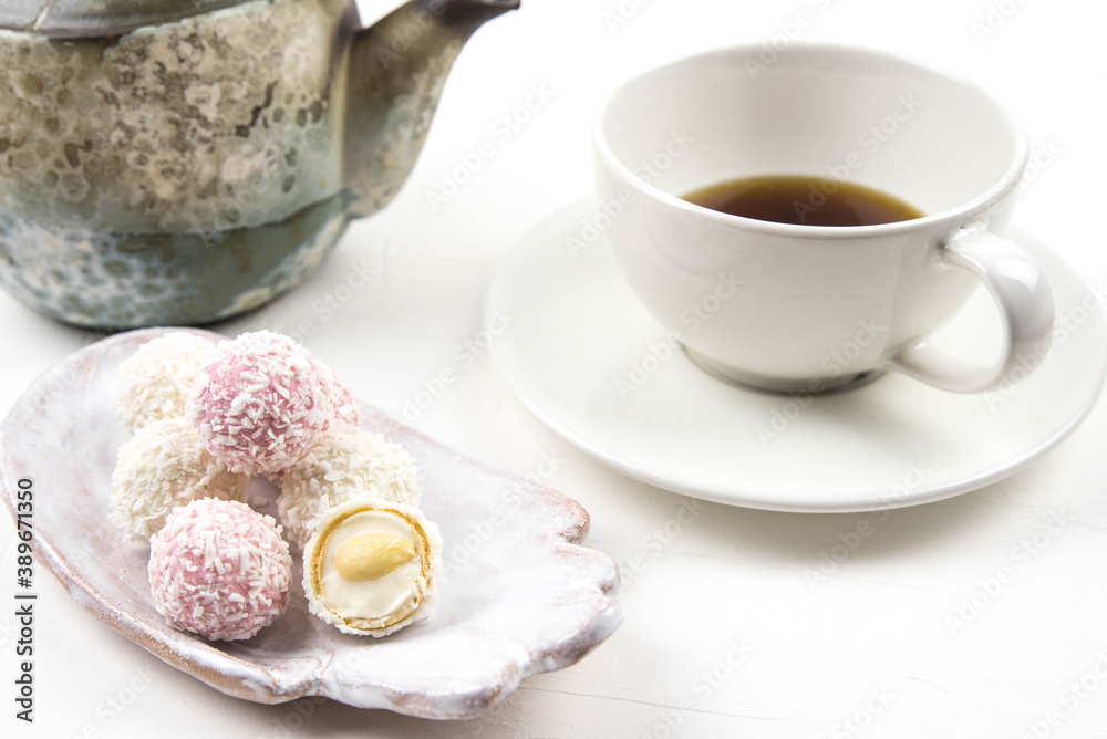 White coconut candy balls on handmade plate. Coconut cookies on white background with the cup of tea. Teapot with black tea. White coconut candy balls on handmade plate. Coconut cookies on white background with the cup of tea. Teapot with black tea.
