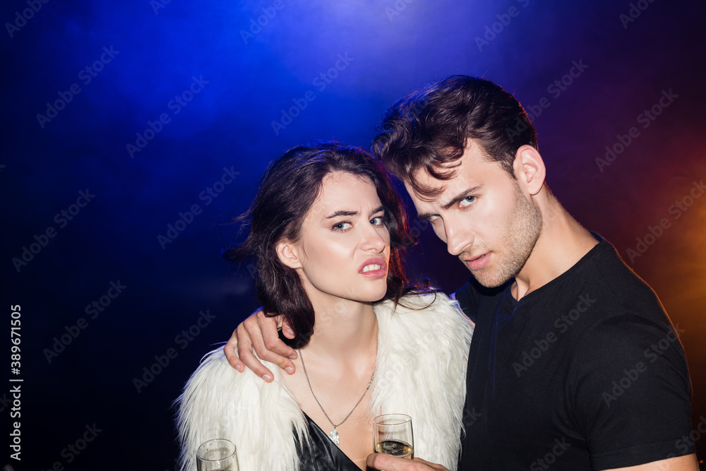 Irritated couple with glasses of champagne hugging and looking at camera with backlit on black Irritated couple with glasses of champagne hugging and looking at camera with backlit on black