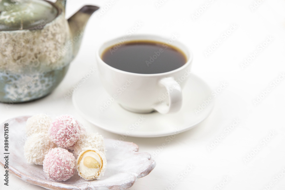 White coconut candy balls on handmade plate. Coconut cookies on white background with the cup of tea. Teapot with black tea. 