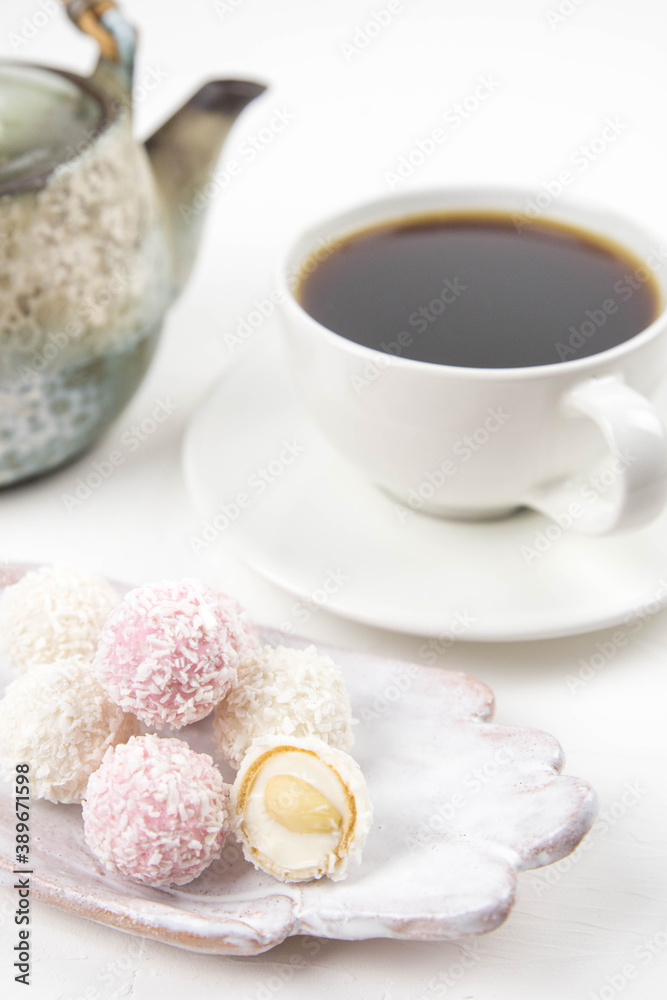White coconut candy balls on handmade plate. Coconut cookies on white background with the cup of tea. Teapot with black tea. White coconut candy balls on handmade plate. Coconut cookies on white background with the cup of tea. Teapot with black tea.