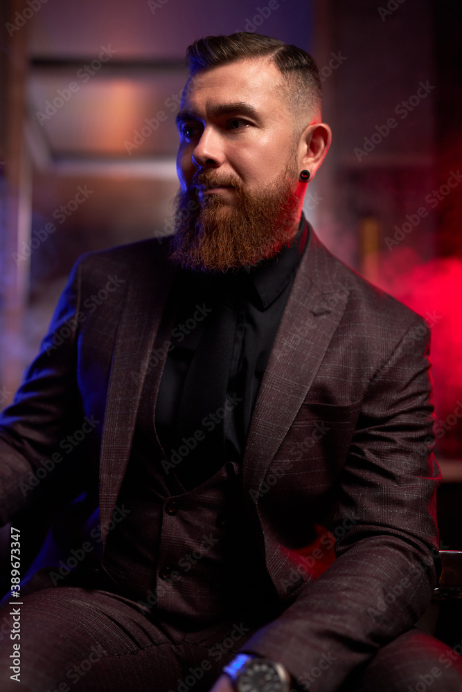 handsome young bearded man in a tuxedo sitting in dark room in fashionable clothing for the festive evening