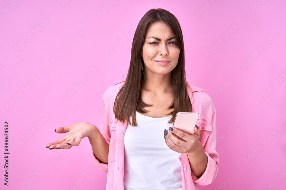Brunette attractive girl holding mobile phone in hand with puzzled face and thinking, isolated in pink studio with copy space Brunette attractive girl holding mobile phone in hand with puzzled face and thinking, isolated in pink studio with copy space
