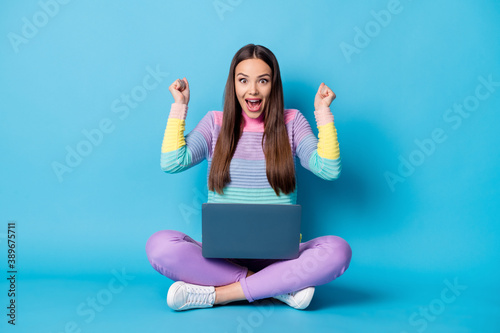 Portrait of pretty glad cheerful girl sitting lotus position crossed legs using laptop having fun isolated over bright blue color background © deagreez