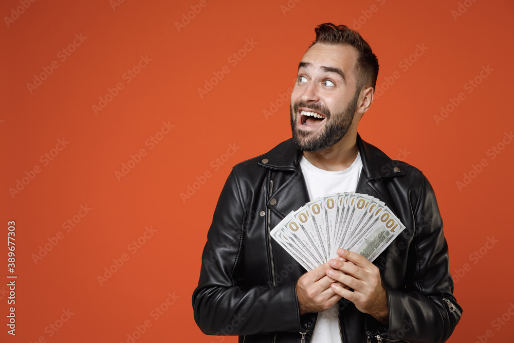 Excited young bearded man in basic white t-shirt, black leather jacket standing hold fan of cash money in dollar banknotes looking aside isolated on bright orange colour background studio portrait. Excited young bearded man in basic white t-shirt, black leather jacket standing hold fan of cash money in dollar banknotes looking aside isolated on bright orange colour background studio portrait.