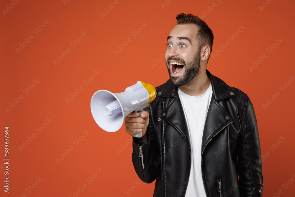 Surprised young bearded man 20s wearing casual basic white t-shirt, black leather jacket standing screaming in megaphone looking aside isolated on bright orange colour background studio portrait. Surprised young bearded man 20s wearing casual basic white t-shirt, black leather jacket standing screaming in megaphone looking aside isolated on bright orange colour background studio portrait.