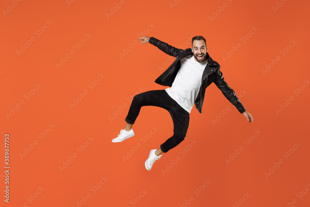 Full length of excited cheerful young bearded man 20s in basic white t-shirt black leather jacket jumping spreading hands looking camera isolated on bright orange colour background studio portrait.
