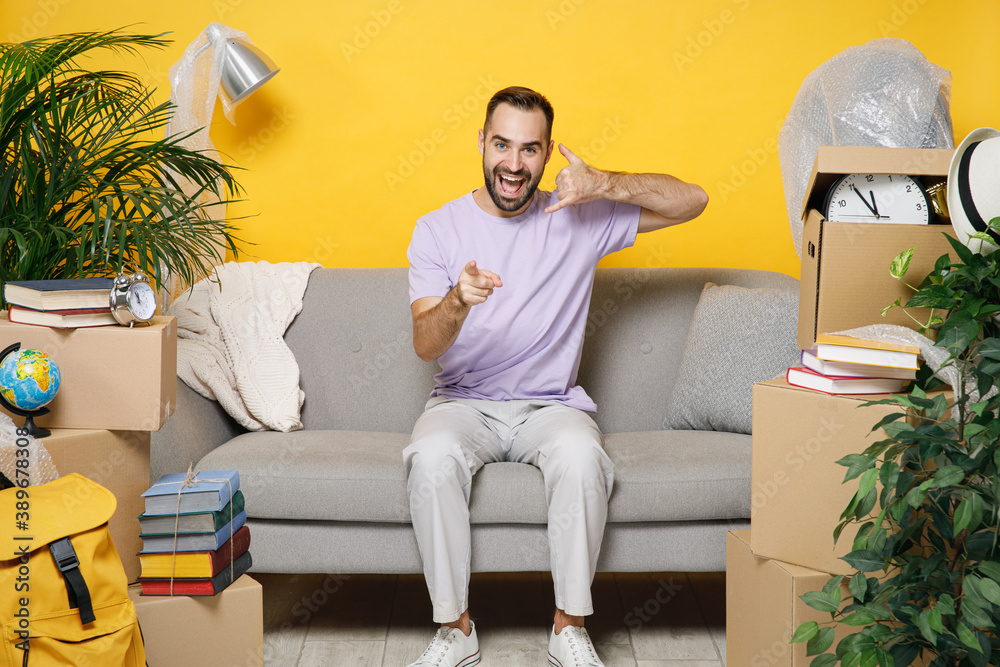 Excited owner man doing phone gesture call me point on camera sits in living room on sofa at home household unpacking stuff rents flat isolated on yellow wall. Relocation moving in apartment concept. Excited owner man doing phone gesture call me point on camera sits in living room on sofa at home household unpacking stuff rents flat isolated on yellow wall. Relocation moving in apartment concept.