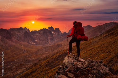 Woman Standing on Rocks looking at Scenic Mountain Peaks and Valley during Fall in Canadian Nature. Dramatic Twilight Sky Artistic Render. Landscape in Tombstone Territorial Park, Yukon, Canada.