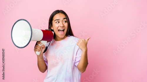 Young asian woman holding a megaphone isolated on pink background points with thumb finger away, laughing and carefree.