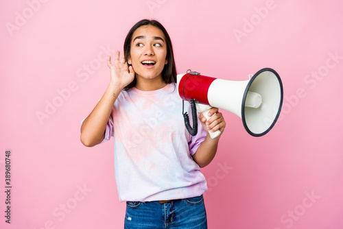Young asian woman holding a megaphone isolated on pink background trying to listening a gossip.
