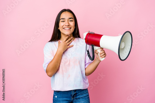 Young asian woman holding a megaphone isolated on pink background laughs out loudly keeping hand on chest.