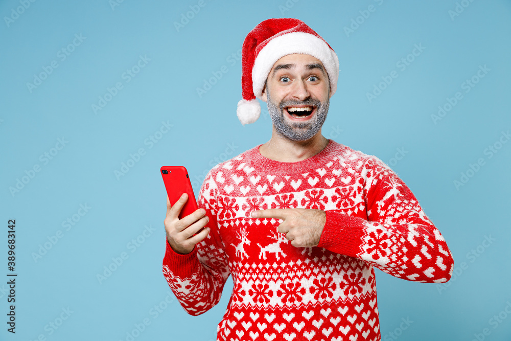 Excited Northern bearded man frozen snow face in Santa hat Christmas sweater pointing index finger on mobile phone isolated on blue background. Happy New Year celebration holiday winter time concept. Excited Northern bearded man frozen snow face in Santa hat Christmas sweater pointing index finger on mobile phone isolated on blue background. Happy New Year celebration holiday winter time concept.