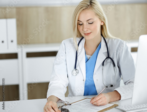 Woman-doctor at work while sitting at the desk in hospital or clinic. Blonde cheerful physician filling up medication history record form