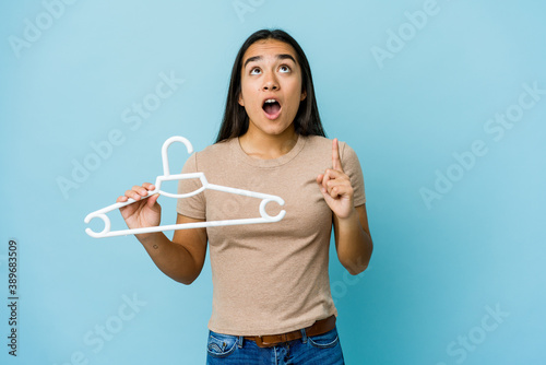 Young asian woman holding a hanger isolated on blue background pointing upside with opened mouth.