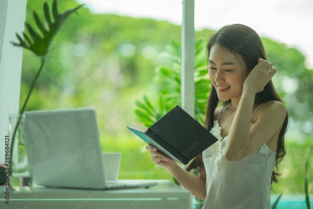 Beautiful young Asian woman in cozy pajamas reading a book while resting on the window sill at home. Beautiful young Asian woman in cozy pajamas reading a book while resting on the window sill at home.