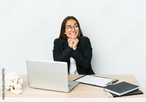 Young traumatologist asian woman isolated on white background keeps hands under chin, is looking happily aside.